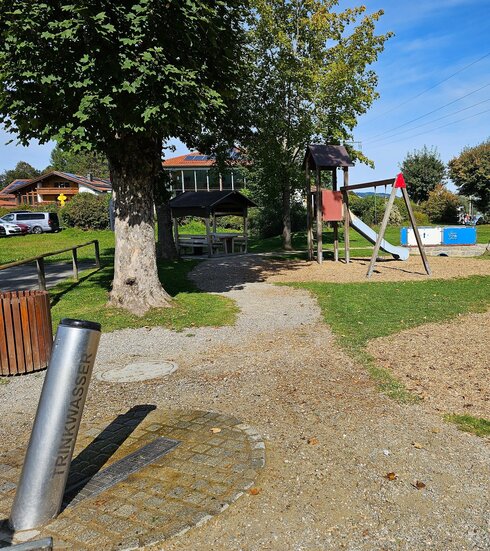 Das Bild zeigt einen Trinkbrunnen bestehend aus einem langen Metallrohr. Darauf ist „Trinkwasser“ geschrieben. Der Trinkbrunnen steht am Rand eine s Spielplatzes. Eine Schaukel, Rutsche und weitere Spielgeräte sind zu sehen. Vereinzelt wachsen hohe Bäume um und auf dem Spielplatz. Der Himmel ist blau. Die Sonne scheint