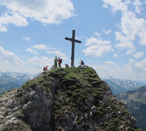 Das Bild zeigt ein Gipfelkreuz, auf dem mehrere Personen sitzen und stehen und in die Ferne blicken. Im Hintergrund sind unzählige Gipfel zu sehen. Der Himmel ist blau. Die Sonne scheint.    