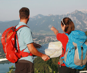 Das Bild fängt einen Moment der Abenteuer und Erkundung ein. Zwei Personen, einem Mann und einer Frau, stehen auf einem Berg, ihre Rücken zeigen zur Kamera. Sie sind beide mit Rucksäcken ausgestattet, was darauf hinweist, dass sie auf einem Hike oder Trek sind. Der Mann deutet auf eine Karte. Die Frau deutet in die Ferne. Der Hintergrund sieht man über eine Ebene aus Dörfern, See und Natur. Der Himmel über ihnen ist blau und klar.