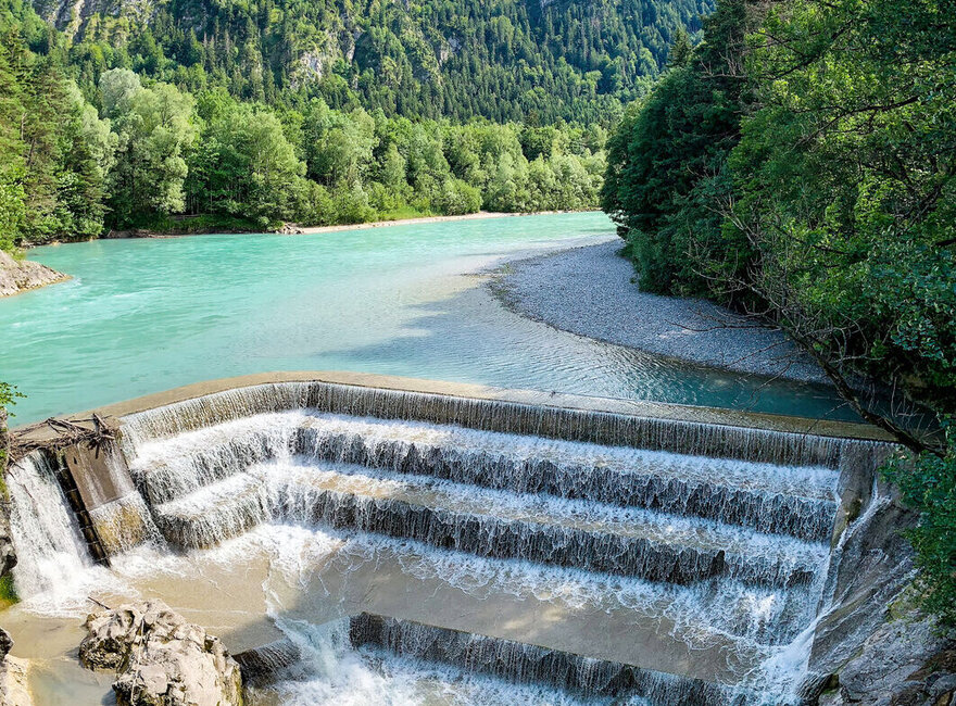 Das Bild zeigt einen künstilich angelegten Wasserfall, den Lechfall in Füssen. Im Hintergreund ist der Fluss Lech und Bäume im Sonnenschein unter blauem Himmel zu erkennen. 