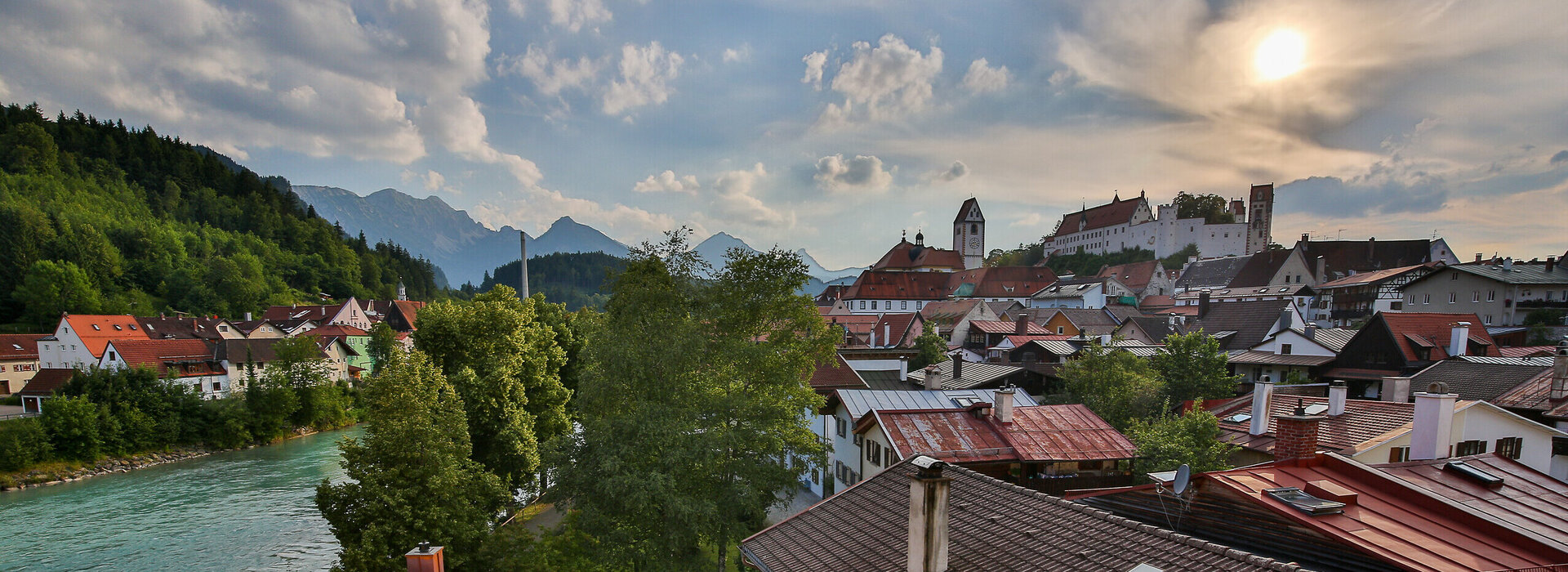 Das Foto zeigt im Hintergrund das Hohe Schloss, das auf einer Anhöhe über Füssen thront. Davor sieht man über die roten Dächer von Füssen. Am linken Bildrand läuft der türkisblaue Fluss Lech. Dahinter erheben sich die Berge. Der Himmel ist blau mit wenigen weißen Wolken durchzogen. Die Abendsonne taucht die Szene in ein warmes Licht. 