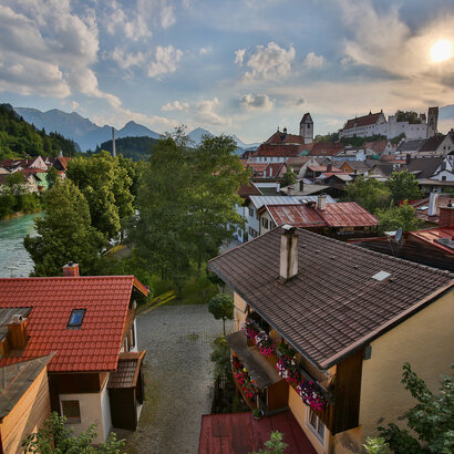Das Foto zeigt im Hintergrund das Hohe Schloss, das auf einer Anhöhe über Füssen thront. Davor sieht man über die roten Dächer von Füssen. Am linken Bildrand läuft der türkisblaue Fluss Lech. Dahinter erheben sich die Berge. Der Himmel ist blau mit wenigen weißen Wolken durchzogen. Die Abendsonne taucht die Szene in ein warmes Licht. 