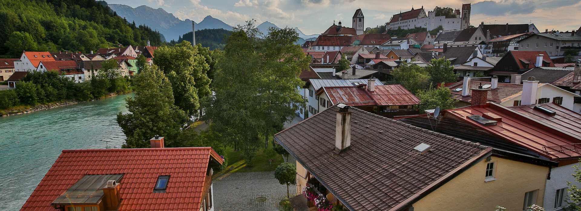 Das Foto zeigt im Hintergrund das Hohe Schloss, das auf einer Anhöhe über Füssen thront. Davor sieht man über die roten Dächer von Füssen. Am linken Bildrand läuft der türkisblaue Fluss Lech. Dahinter erheben sich die Berge. Der Himmel ist blau mit wenigen weißen Wolken durchzogen. Die Abendsonne taucht die Szene in ein warmes Licht. 