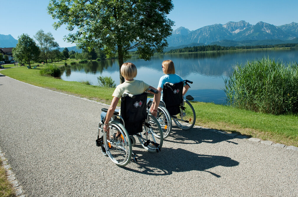 aziergang entlang eines gepflasterten Weges genießen. Der Weg, der aus grauviolettem Kies besteht, ist von grünem Laub bedeckt und bietet ein natürliches und friedliches Kulisse. Die Frauen, beide in legere Kleidung gekleidet, sitzen bequem in ihren Rollstühlen, die mit Handbremsen für zusätzliche Sicherheit ausgestattet sind. Der Weg führt sie zu einem friedlichen See, in dessen ruhiges Wasser der blaue Himmel darüber spiegelt. Die Atmosphäre des Bildes ist eine von Entspannung und Genuss der Natur.