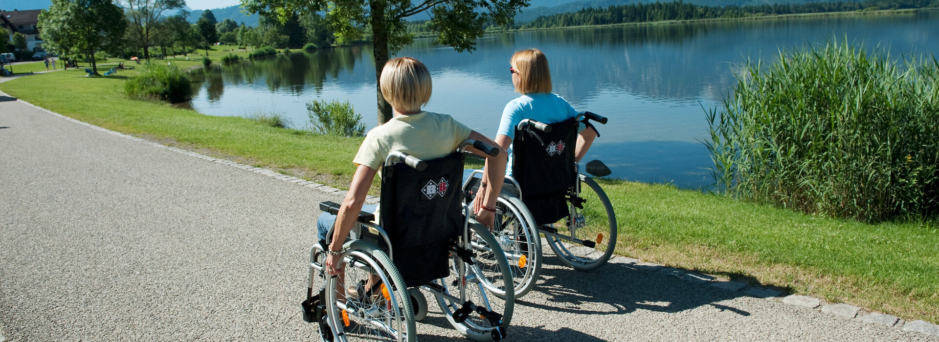 aziergang entlang eines gepflasterten Weges genießen. Der Weg, der aus grauviolettem Kies besteht, ist von grünem Laub bedeckt und bietet ein natürliches und friedliches Kulisse. Die Frauen, beide in legere Kleidung gekleidet, sitzen bequem in ihren Rollstühlen, die mit Handbremsen für zusätzliche Sicherheit ausgestattet sind. Der Weg führt sie zu einem friedlichen See, in dessen ruhiges Wasser der blaue Himmel darüber spiegelt. Die Atmosphäre des Bildes ist eine von Entspannung und Genuss der Natur.