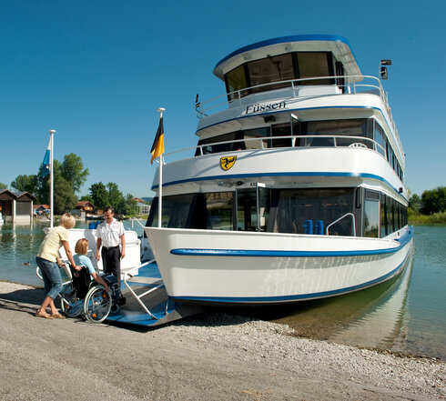 Das Foto zeigt eine sanfte Szene in einer Marina. Ein großes, weißes Motorschiff, das mit Blautönen und gelben Akzenten versehen ist, liegt am Pier. Das Schiff ist der Mittelpunkt des Bildes und steht wegen seiner Größe und seiner Farbe hervor.  Am Pier versammeln sich verschiedene Menschen um das Schiff. Sie scheinen das Schiff zu bestaunen oder möglicherweise in Gedanken versunken über eine Wasserfahrt nachzudenken. Unter ihnen befindet sich auch eine Person im Rollstuhl, die der Szene eine Prise Diversität verleiht.  Die Marina selbst befindet sich inmitten üppiger Vegetation, mit Bäumen und Sträuchern, die den Hintergrund bilden. Das ruhige Wasser des Sees spiegelt die Ruhe der Szene wider. Der Himmel darüber ist ein kräftiges Blau, das auf einen wunderschönen Tag hindeutet.  Insgesamt ergibt das Bild das Bild eines genüsslichen Ausflugs, mit dem Schiff als Symbol für Abenteuer und Erkundung.