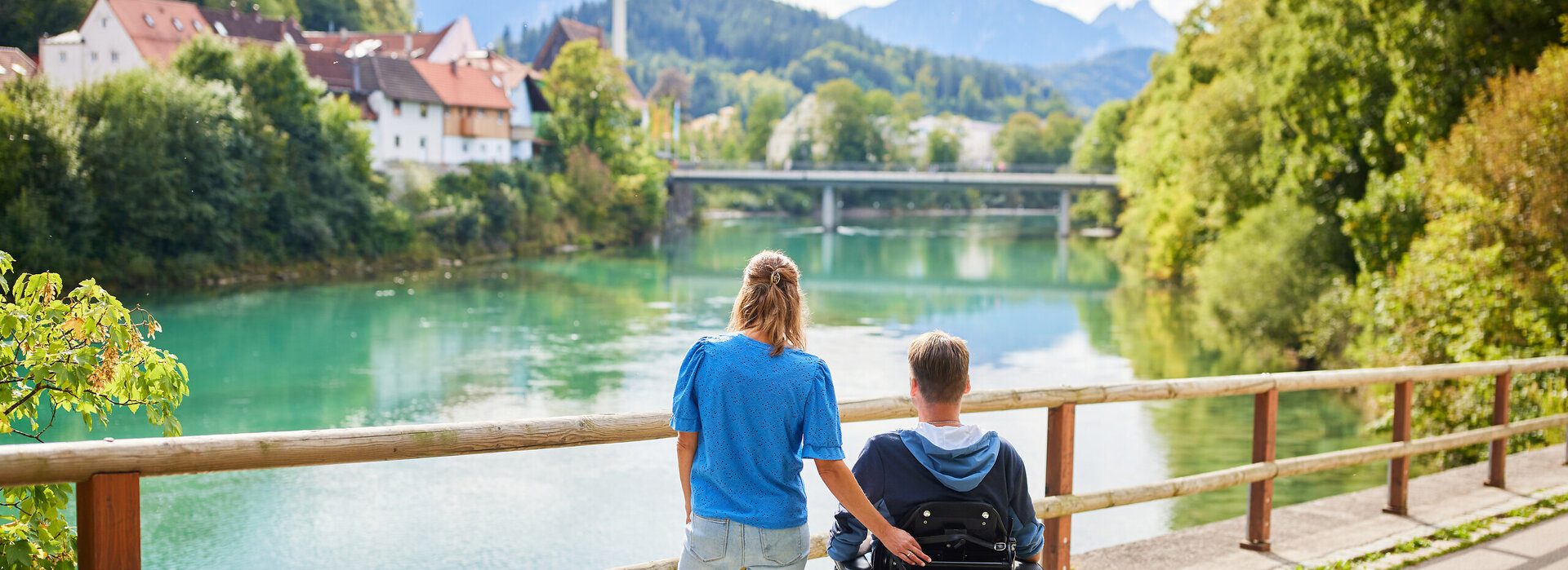 Das Bild fängt einen friedlichen Moment über einer Holzbrücke ein, die einen Blick auf eine Flusslandschaft bietet. Zwei Personen, ein Mann und eine Frau, stehen auf der Brücke, mit dem Rücken zur Kamera. Der Mann sitzt im Rollstuhl, während die Frau neben ihm steht. Beide sind lässig gekleidet, wobei die Frau ein blaues Shirt und der Mann einen schwarzen Mantel trägt. Die Brücke aus Holzdielen spannt sich über den Fluss und bietet einen Aussichtspunkt für den Betrachter. Der Fluss selbst ist ein lebendiges Blau, das den klaren Himmel darüber widerspiegelt. Im Hintergrund schmiegt sich ein malerisches Dorf in die üppige Vegetation, der das Bild mit Charme erfüllt. Die Atmosphäre des Bildes ist eine der Ruhe und Gesellschaft.