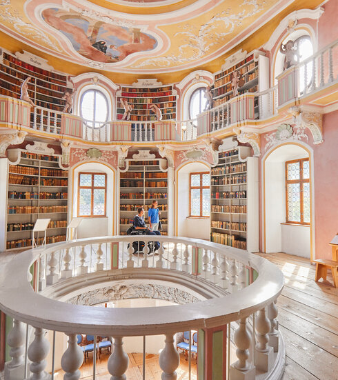 Das Bild zeigt eine imposante, kreisrunde Bibliothek mit einer hohen Decke. Die Wände sind in einem warmen, rosafarbenen Ton gemalt, und die Decke ist mit aufwendigen Mustern und wunderschönen Verzierungen geschmückt.