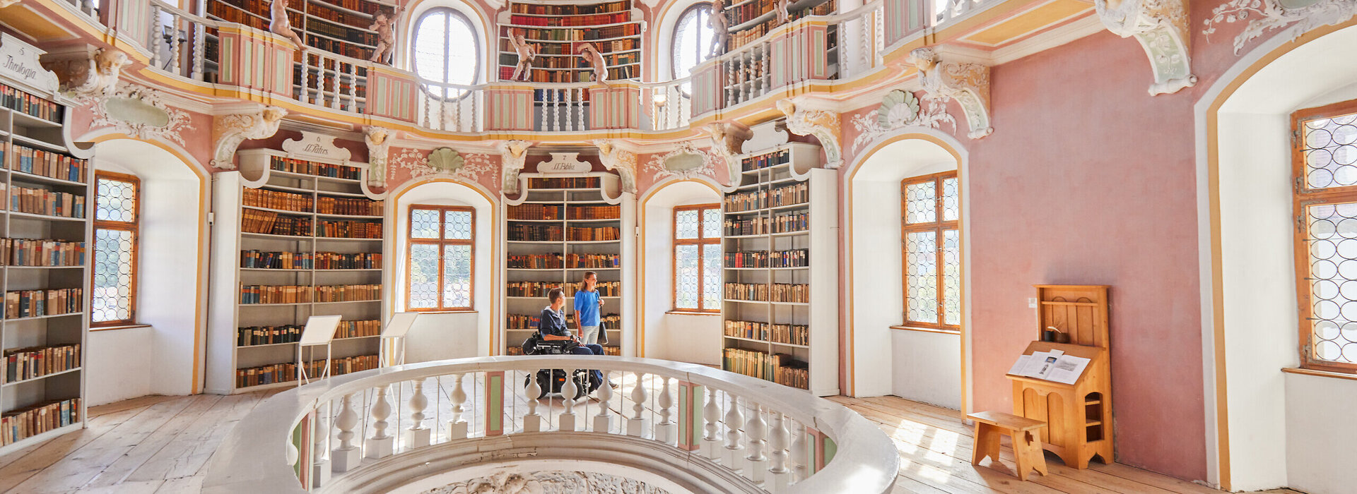 Das Bild zeigt eine imposante, kreisrunde Bibliothek mit einer hohen Decke. Die Wände sind in einem warmen, rosafarbenen Ton gemalt, und die Decke ist mit aufwendigen Mustern und wunderschönen Verzierungen geschmückt.