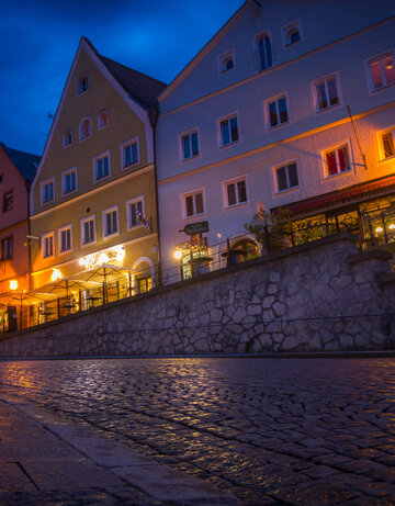 Das Bild zeigt eine herzige europäische Straße bei Nacht, im warmen Glanz der Straßenlampen. Die Straße ist von bunten Gebäuden gesäumt, deren Fassaden mit zahlreichen Fenstern geschmückt sind. Die Gebäude, in Tönen von Gelb, Orange und Weiß gestrichen, stehen hoch und widerspiegelnd der Straße, was eine idyllische Szene schafft. Die Kopfsteinpflasterstraße, rutschig mit dem Nachtregen, fügt dem Geschehen einen Hauch von rustikalem Charme hinzu.