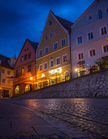 Das Bild zeigt eine herzige europäische Straße bei Nacht, im warmen Glanz der Straßenlampen. Die Straße ist von bunten Gebäuden gesäumt, deren Fassaden mit zahlreichen Fenstern geschmückt sind. Die Gebäude, in Tönen von Gelb, Orange und Weiß gestrichen, stehen hoch und widerspiegelnd der Straße, was eine idyllische Szene schafft. Die Kopfsteinpflasterstraße, rutschig mit dem Nachtregen, fügt dem Geschehen einen Hauch von rustikalem Charme hinzu.