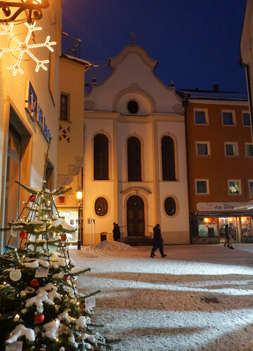 Das Bild fängt eine friedliche Nachtszene in einer europäischen Stadt ein. Der Fokus liegt auf einem wunderschön dekorierten Weihnachtsbaum, der in der Vordergrund steht. Der Baum ist mit funkelndem Lichtern und Ornamenten geschmückt, die eine warme Glut auf die umgebende Fläche werfen.  Im Hintergrund steht eine große Kirche mit einer weißen Fassade und einem roten Dach majestätisch. Die architektonischen Details der Kirche sind deutlich sichtbar und fügen dem Charme der Szene hinzu.  Die Straße, wo der Baum steht, ist von Gebäuden gesäumt, deren Fenster mit dem weichen Licht der Kirche leuchten. Einige Menschen können gesehen werden, die auf der Straße gehen, fügen einen Hauch von Leben zu dem sonst stillen Bild hinzu.  Der Himmel über ist ein tiefes Blau, das eine eindrucksvolle Kulisse für die festlichen Dekorationen bietet. Die allgemeine Atmosphäre des Bildes ist eine der Ruhe und Gelassenheit, die die Magie der Weihnachtszeit einfängt.