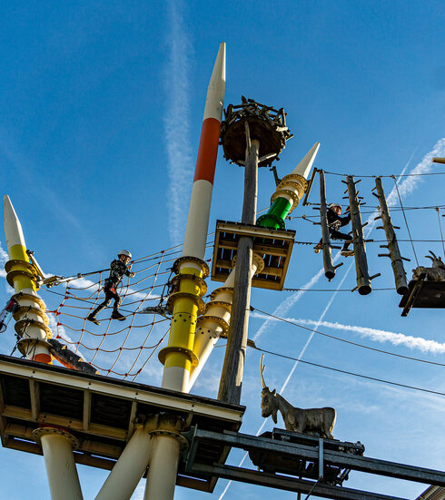 Das Foto zeigt einen Teil eines Kletterparks, dessen Stangen wie Mikado Stäbe lackiert sind. Zwischen den Seilen und Netzen, die die unterschiedlichen Stangen verbinden, sind Trittstufen und Plattformen unterschiedlicher Größe angebracht. Dort klettern ein Mädchen und ein Junge mit Helmen und Kletterausrüstung gesichert. Der Himmel ist blau mit wenigen weißen Wolken durchzogen.