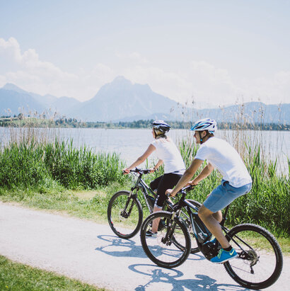 Das Bild zeigt einen Mann und eine Frau, die Fahrrad auf einem Fahrradweg entlang des Hopfensees fahren. Der Fahrradweg ist führt an grünen Wiesen entlang. Im Hintergrund sieht man den Hopfensee und die Berge. Die Radfahrer sind in lässiger Kleidung und mit Helm gekleidet. Beide tragen ein weißes T-Shirt und eine kurze Hose. Das Bild vermittelt eine entspannte Fahrradfahrt.