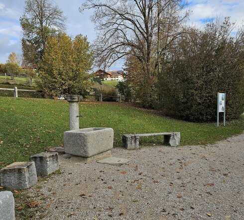 Man sieht eine Platz, der mit Steinquadern eingerahmt ist. Die Steinquader bieten kleine Sitzgelegenheiten. Zwischen den Steinquadern steht ein steinerner Brunnen. Der Boden des Platzes ist mit Kies bedeckt. Der Platz grenzt an eine Wiese, die einen kleinen Hang nach oben läuft und durch Büsche, Bäume und einen Holzzaun begrenzt ist. Im Hintergrund sind mehrere Häuser auf einer Anhöhe zu sehen. Der Himmel ist blau und mit weißen Wolken durchzogen. 