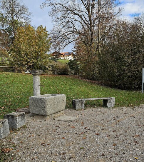 Man sieht eine Platz, der mit Steinquadern eingerahmt ist. Die Steinquader bieten kleine Sitzgelegenheiten. Zwischen den Steinquadern steht ein steinerner Brunnen. Der Boden des Platzes ist mit Kies bedeckt. Der Platz grenzt an eine Wiese, die einen kleinen Hang nach oben läuft und durch Büsche, Bäume und einen Holzzaun begrenzt ist. Im Hintergrund sind mehrere Häuser auf einer Anhöhe zu sehen. Der Himmel ist blau und mit weißen Wolken durchzogen. 