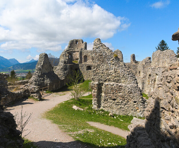 Das Bild zeigt die Mauerreste einer Burg. Die Mauern sind teilweise noch gut erhalten und reichen über zwei Stockwerke. Innerhalb der Burgruine verlaufen Wege. Es wächst aber auch Gras und Sträucher. Im Hintergrund erheben sich Berge und grün, bewaldete Hügel. Der Himmel ist blau. Die Sonne scheint. 