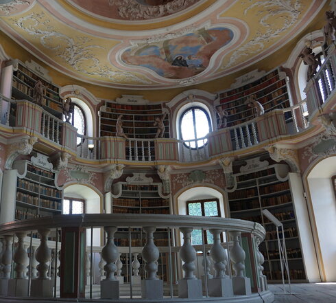 Das Foto zeigt den runden Raum einer Bibliothek. Der Raum ist mit Bücherregalen gefüllt, die nur von Fenstern unterbrochen werden. Die Wände sind in altrosa gestrichen. Der Raum erstreckt sich über zwei Etagen. Im Bereich der oberen Etage verläuft eine Balustrade ringsum. Auf dem Geländer der Balustrade sind im Abstand von ca. 1 Meter unterschiedliche Putten und Engel platziert. Der Raum öffnet sich über eine ovale Öffnung auch in den Raum darunter. Um die Öffnung herum ist ebenfalls ein weißes Geländer angebracht. Die Decke des Raums ist mit Stuck und Gemälden verziert. 