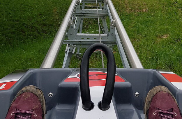 Das Bild zeigt einen fröhlichen Moment eines Vaters und eines Sohnes auf einer Wasserrutsche. Der Vater, bekleidet mit einem blauen Hemd, sitzt dem Sohn gegenüber, der ein grünes Hemd trägt. Beide halten sich an den Rand der Rutsche, ihre Gesichter erstrahlen vor Aufregung. Die Rutsche besteht aus blauem Kunststoff und ist in einem üppigen Grünfeld aufgestellt, umgeben von Bäumen und Buschwerk. Der Vater und der Sohn sind der Hauptraum des Bildes, ihre bunten Hemden stehen im Kontrast zum natürlichen Hintergrund hervor. Das Bild vermittelt eine Atmosphäre von Spaß und Abenteuer, wenn die beiden ihre Zeit auf der Rutsche genießen.