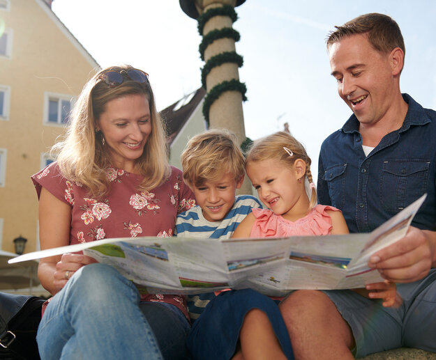  Das Bild ist ein Farbfoto, das eine Familie von vier Personen zeigt, die draußen sitzen. Die Frau, die wahrscheinlich die Mutter ist, ist auf der linken Seite des Bildes zu sehen, sie sitzt und hält eine Zeitung aufgeschlagen vor sich. Sie lächelt und sieht in die Kamera. Neben ihr sitzt ein junges Mädchen, ebenfalls lächelnd und in die Kamera blickend. Das Mädchen trägt ein Rosa-Top. Auf der rechten Seite des Bildes sitzt ein Mann, der ebenfalls die Zeitung aufgeschlagen vor sich hält. Er lächelt und sieht in die Kamera. Der Mann trägt ein Blau-Shirt. Im Hintergrund ist ein gelbes Gebäude mit mehreren Fenstern zu sehen. Der Himmel ist klar und blau, was auf einen sonnigen Tag hindeutet. Es sind keine sichtbaren Texte oder Logos im Bild zu sehen.