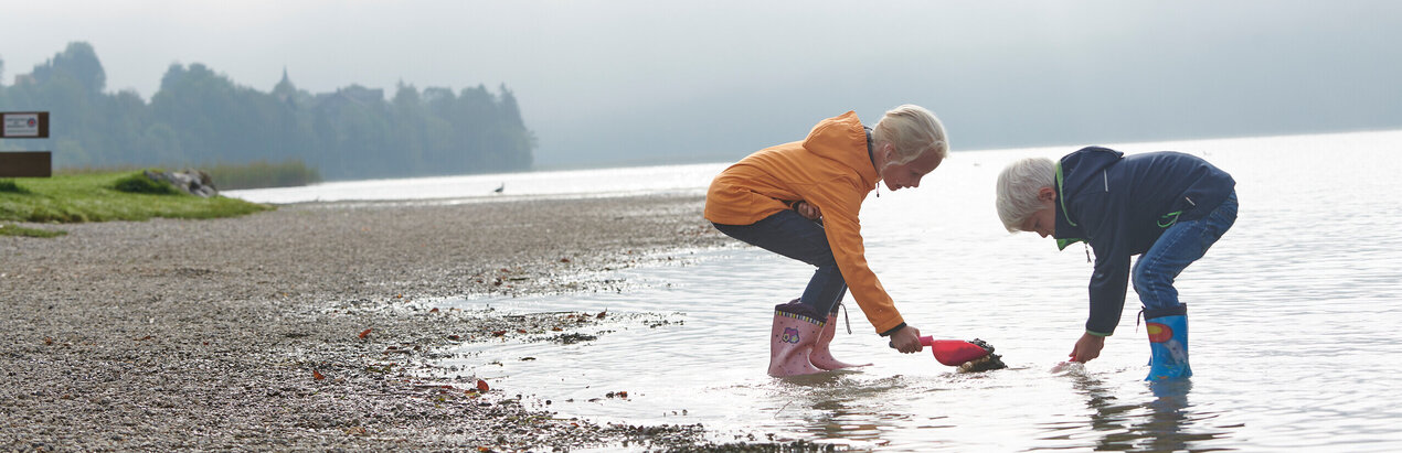 Das Bild zeigt einen Jungen und ein Mädchen. Sie stehen am flachen Seeufer im knöchelhohen Wasser. Beide tragen Regenjacken und Gummistiefel. Das Wetter ist nebelig. 