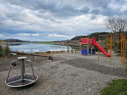Auf dem Bild sieht man einen Spielplatz, der an eienem Seeufer liegt. Es ist eine rote Rutsche und ein Karusell zu sehen.