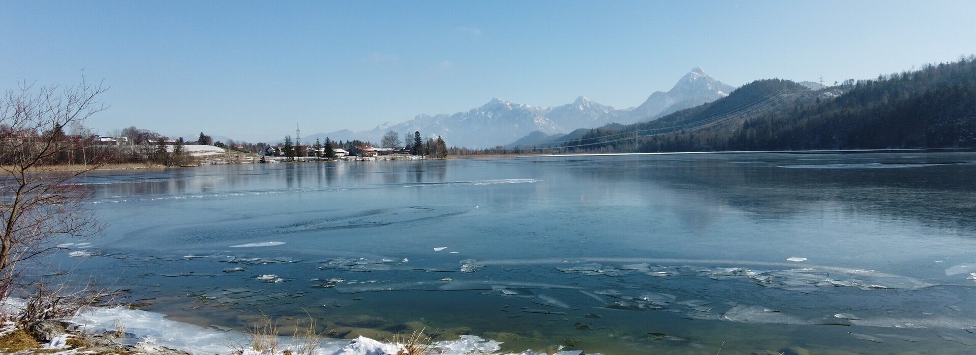 Das Bild zeigt einen See, dessen Wasseroberfläche zugefroren ist. Die Eisschicht liegt an einigen Stellen wie in Scherben. Im Hintergrund erheben sich die Berge und bewaldeten Hügel. Der Himmel ist blau. Die Sonne scheint. 
