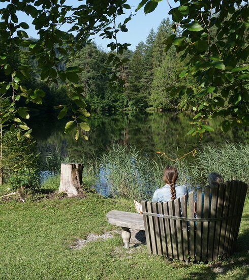 Das Bild zeigt im Vordergrund eine hölzerne Bank mit Rückenlehne, die sich auf einer grünen Wiese befindet. Eine Frau sitzt mit dem Rücken zur Kamera auf der Bank, ihre Haare sind zu einem Zopf gebunden. Sie scheint auf den vor ihr liegenden See, der von Bäumen und Vegetation umgeben ist, zu blicken. Das Wasser reflektiert die Vegetation und schafft eine beruhigende Atmosphäre. Der Himmel ist blau. 