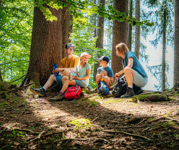 Das Bild zeigt eine Familie, bestehend aus einer Frau einem Mann, einem Mädchen und einem Jungen. Sie tragen sportliche Freizeitkleidung. Sie sitzen auf dem Waldboden. Rings um sie herum wachsen Bäume. Sie halten Trinkflaschen in den Händen und lächeln sich zu. Der Himmel ist blau. Die Sonne scheint durch die Äste und Zweige der Bäume. 