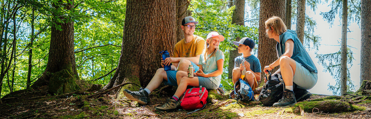 Das Bild zeigt eine Familie, bestehend aus einer Frau einem Mann, einem Mädchen und einem Jungen. Sie tragen sportliche Freizeitkleidung. Sie sitzen auf dem Waldboden. Rings um sie herum wachsen Bäume. Sie halten Trinkflaschen in den Händen und lächeln sich zu. Der Himmel ist blau. Die Sonne scheint durch die Äste und Zweige der Bäume. 