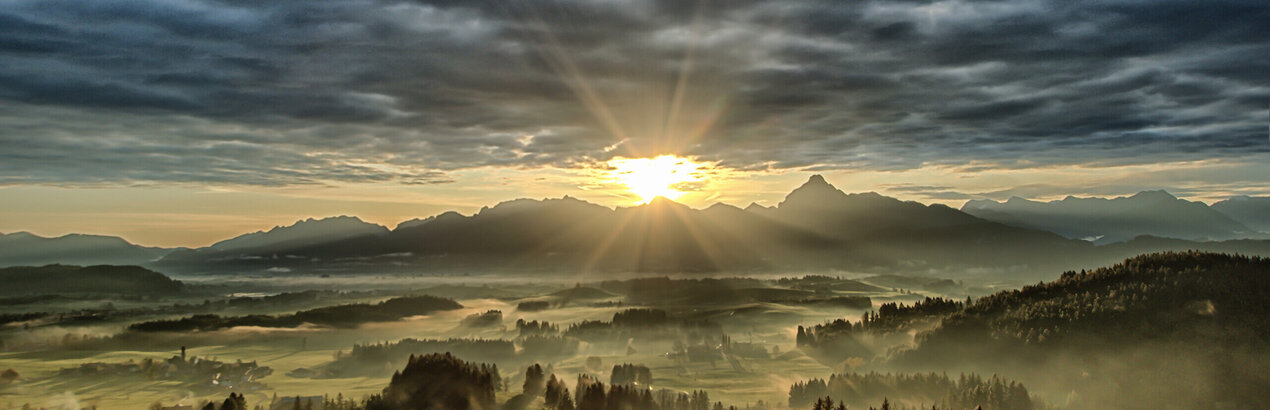 Das Bild zeigt eine atemberaubende Landschaft, unter einem dramatischen Himmel. Der Himmel ist voller dunkler Wolken, die eine Kulisse für die Szene schaffen. Die Sonne scheint hell durch die Wolken und gibt der Landschaft darunter ein warmes Glühen. Die Berge in der Ferne sind majestätisch, mit ihren Spitzen streben sie zum Himmel. Die Vordergrund ist voller grüner Bäume, die einen Farbtupfer in die ansonsten monochrome Szene bringen. Die Gesamtzusammenstellung des Bildes erzeugt ein Gefühl von Ehrfurcht und Bewunderung für die Schönheit der Natur.