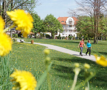 Auf einem befestigten Weg laufen zwei Frauen, die sommerlich gekleidet sind. Der Weg führt durch eine Wiese, auf der auch vereinzelt hohe Bäume wachsen. Der Himmel ist blau. Die Sonne scheint. 
