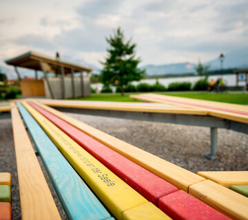 Das Bild zeigt zwei bunte Holzlatten, die in unterschiedlichen Winkeln aufeinander zulaufen. Auf einer gelben Holzlatte ist etwas eingraviert. Mann kann nur „sind an der Seele“ lesen. Im Hintergrund ist ein See zu sehen. Dahinter erheben sich hohe Berge. Der Himmel ist wolkenverhangen. 