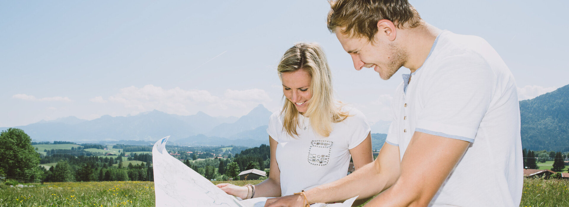 Das Bild zeigt einen Mann und eine Frau, die neben ihren Rädern stehen und eine große Karte lesen. Beide lächeln. Der Mann zeigt auf etwas auf der Karte. Im Hintergrund ist ein blühendes Feld zu sehen. Dahinter erheben sich Berge und bewaldete Hügel. Der Himmel ist blau. Die Sonne scheint.  