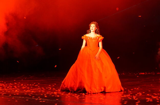 Das Foto zeigt eine Frau in einem historischen roten Kleid mit einem sehr weiten ausladenden, roten Rock, der bis zum Boden reicht. Die Frau hat lange braune Haare. Die Scene ist in rotes Licht getaucht. Am Boden liegen unzählige rote Rosenblätter. Die Szene zeigt die Kaiserin Sisi aus dem Musical Ludwig² - Der König ist zurück.