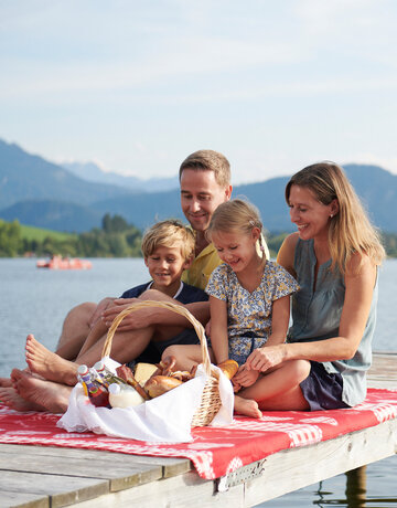 Das Bild fängt einen friedlichen Familienmoment am Ufer eines Sees ein. Eine Familie bestehend aus einem Mann, einer Frau und zwei Kindern sitzt auf einer Holzdiele. Der Mann sitzt auf der linken Seite, die Frau sitzt in der Mitte und die zwei Kinder sitzen auf der rechten Seite. Sie alle lächeln und schauen zur Kamera, wodurch eine warme und glückliche Atmosphäre geschaffen wird.  Die Familie wird von der Natur umgeben, mit dem See und den Bergen im Hintergrund. Die Holzdiele, auf welcher die Familie sitzt, ist rot und weiß, was eine Farbtupfer im Bild schafft.  Im Vordergrund befindet sich ein Korb voller Essen, was darauf hindeutet, dass die Familie vielleicht ein Picknick hat. Die Gesamtszene ist eine der Entspannung und Freude an der Natur.