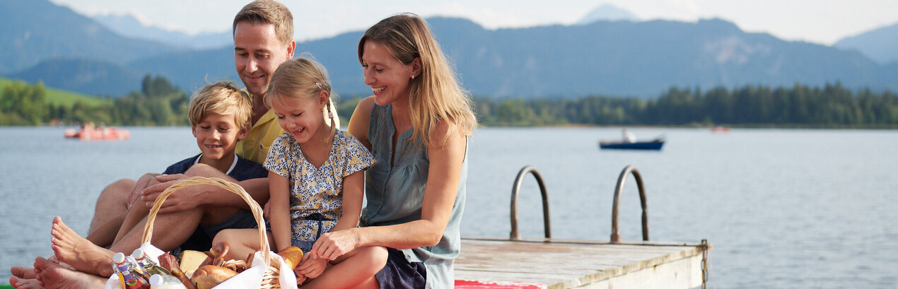 Das Bild fängt einen friedlichen Familienmoment am Ufer eines Sees ein. Eine Familie bestehend aus einem Mann, einer Frau und zwei Kindern sitzt auf einer Holzdiele. Der Mann sitzt auf der linken Seite, die Frau sitzt in der Mitte und die zwei Kinder sitzen auf der rechten Seite. Sie alle lächeln und schauen zur Kamera, wodurch eine warme und glückliche Atmosphäre geschaffen wird.  Die Familie wird von der Natur umgeben, mit dem See und den Bergen im Hintergrund. Die Holzdiele, auf welcher die Familie sitzt, ist rot und weiß, was eine Farbtupfer im Bild schafft.  Im Vordergrund befindet sich ein Korb voller Essen, was darauf hindeutet, dass die Familie vielleicht ein Picknick hat. Die Gesamtszene ist eine der Entspannung und Freude an der Natur.