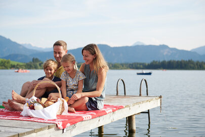 Das Bild fängt einen friedlichen Familienmoment am Ufer eines Sees ein. Eine Familie bestehend aus einem Mann, einer Frau und zwei Kindern sitzt auf einer Holzdiele. Der Mann sitzt auf der linken Seite, die Frau sitzt in der Mitte und die zwei Kinder sitzen auf der rechten Seite. Sie alle lächeln und schauen zur Kamera, wodurch eine warme und glückliche Atmosphäre geschaffen wird.  Die Familie wird von der Natur umgeben, mit dem See und den Bergen im Hintergrund. Die Holzdiele, auf welcher die Familie sitzt, ist rot und weiß, was eine Farbtupfer im Bild schafft.  Im Vordergrund befindet sich ein Korb voller Essen, was darauf hindeutet, dass die Familie vielleicht ein Picknick hat. Die Gesamtszene ist eine der Entspannung und Freude an der Natur.