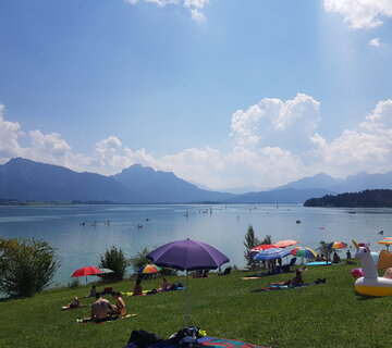 Das Bild zeigt einen See, in dem zahlreiche Menschen schwimmen, plantschen, mit SUP fahren oder am Ufer auf Decken und Handtüchern unter bunten Sonnenschirmen liegen. Der See ist umgeben von Wiesen und Wäldern. Im Hintergrund erheben sich hohe, teilweise bewaldete Berge. Die Sonne scheint. Der Himmel ist blau.