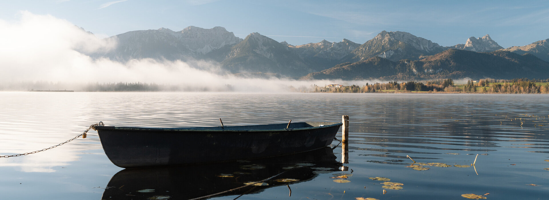 Im Fokus des Bildes steht ein in die Jahre gekommenes Ruderboot, das an einem Holzpfahl befestigt ist und auf einem See schwimmt. Um das Boot herum schwimmen zahlreiche Blätter einer Seerose. Es liegt Nebel über dem See. Im Hintergrund erheben sich die Berge, die noch teilweise vom Nebel verdeckt werden. Der Himmel ist blau. Die Morgensonne scheint.   