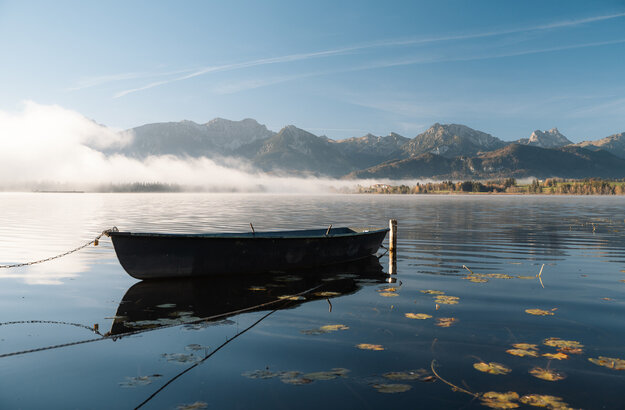 Im Fokus des Bildes steht ein in die Jahre gekommenes Ruderboot, das an einem Holzpfahl befestigt ist und auf einem See schwimmt. Um das Boot herum schwimmen zahlreiche Blätter einer Seerose. Es liegt Nebel über dem See. Im Hintergrund erheben sich die Berge, die noch teilweise vom Nebel verdeckt werden. Der Himmel ist blau. Die Morgensonne scheint.   
