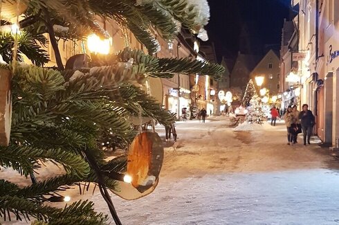 Das Bild fängt eine friedliche Nachtszene in einer Stadt ein. Der Hauptfokus liegt auf einem prächtig geschmückten Weihnachtsbaum, der sich auf der linken Seite des Bildes befindet. Der Baum ist mit weißen Lichtern geschmückt, die der Szene ein festliches Flair verleihen. Der Baum ist von einer schneebedeckten Fläche umgeben, was darauf hinweist, dass es Winter ist.  Im Hintergrund ist eine Straße zu sehen, die von Gebäuden gesäumt wird. Die Straße wird von Straßenlaternen beleuchtet, die auf die Umgebung ein warmes Licht werfen. Einige Menschen können dabei beobachtet werden, wie sie auf der Straße gehen, was ein Gefühl von Leben und Aktivität in die sonst ruhige Szene bringt.  Am Himmel ist es dunkel, was darauf hinweist, dass es Nacht ist. Die allgemeine Atmosphäre des Bildes ist friedlich und festlich und fängt den Charakter einer stillen Winter.