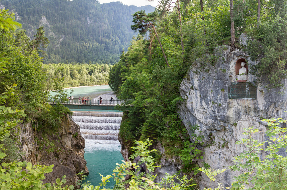 Das Bild fängt einen atemberaubenden Blick auf einen Wasserfall ein, der an einem felsigen Steilhang hinabstürzt. Der Wasserfall liegt im Fokus des Bildes. Das felsige Ufer ist von üppigem grünem Laub und Büschen geziert. Über die Schlucht, die der Wasserfall durchfließt, führt eine Brücke. In einen Felsabhang am Ufer ist eine Büste eingearbeitet. Sie zeigt König Max II. von Bayern. Die Perspektive des Bildes ist aus einer hohen Position, die auf den Wasserfall hinunterblickt, was die Szene zusätzlich in Größe erscheinen lässt.
