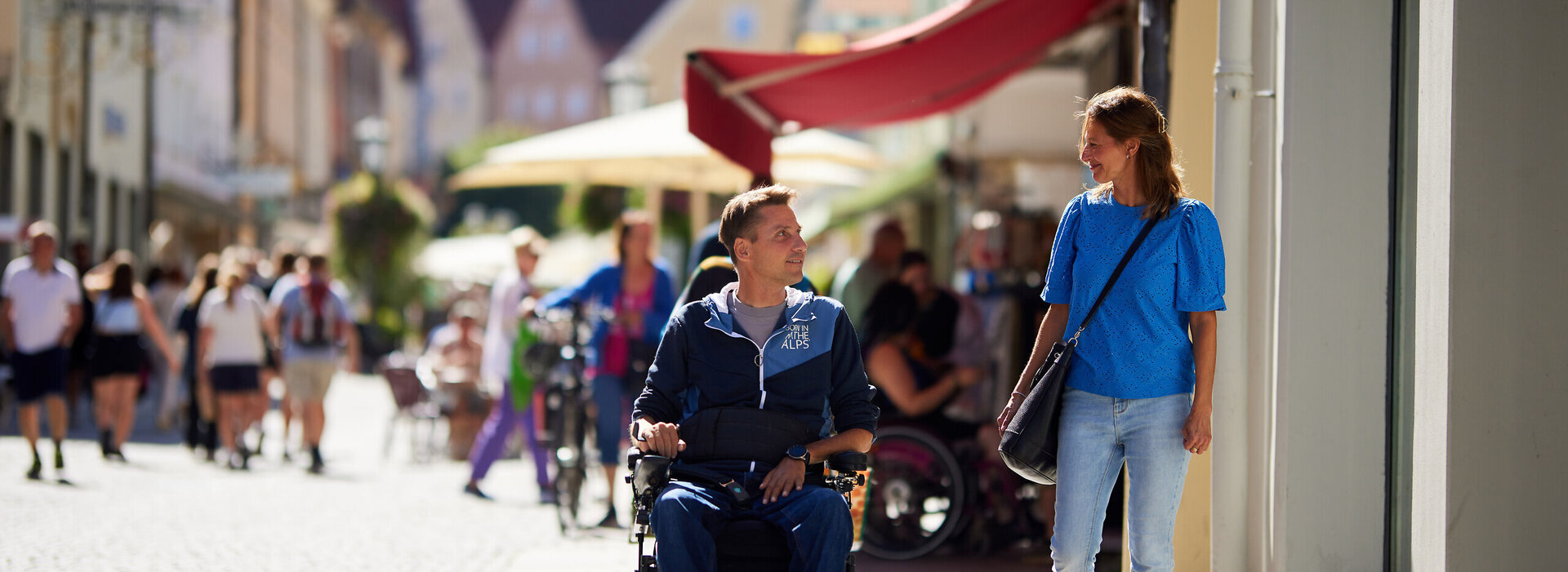  Das Bild zeigt eine lebhafte Straßenszene in einer europäischen Stadt. Der Blickfang des Bildes ist ein Mann im Rollstuhl, der sich in einem Gespräch mit einer Frau befindet, die neben ihm geht. Der Mann ist mit einem blauen Jacket und einem schwarzen Hut gekleidet, während die Frau ein blaues Hemd und blaue Jeans trägt. Sie gehen auf einem Gehweg, der mit unterschiedlichen Geschäften und Restaurants gesäumt ist. Die Straße ist belebt mit Menschen, von denen einige zu Fuß gehen, während andere Fahrräder fahren. Die Gebäude im Hintergrund sind hoch und haben einen europäischen architektonischen Stil. Der Himmel über ihnen ist klar und erlaubt den Schluss auf einen angenehmen Tag. Die Gesamtatmosphäre des Bildes ist lebhaft und voller Leben.