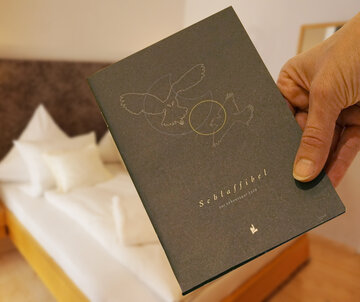 Das Bild zeigt ein dunkelgraues Heft mit der Aufschrift „Schlaffibel“ das eine Hand hält. Im Hintergrund ist unscharf ein weiß bezogenes Doppelbett aus Holz zu sehen.