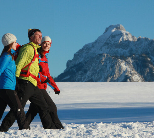 Das Bild zeigt zwei Frauen und einen Mann. Sie tragen Sport-Winterkleidung und laufen nebeneinander auf einem verschneiten Weg. Alle lächeln. Im Hintergrund sieht man die schneebedeckte Umgebung und die verschneiten Berge. Der Himmel ist blau. Die Sonne scheint.  