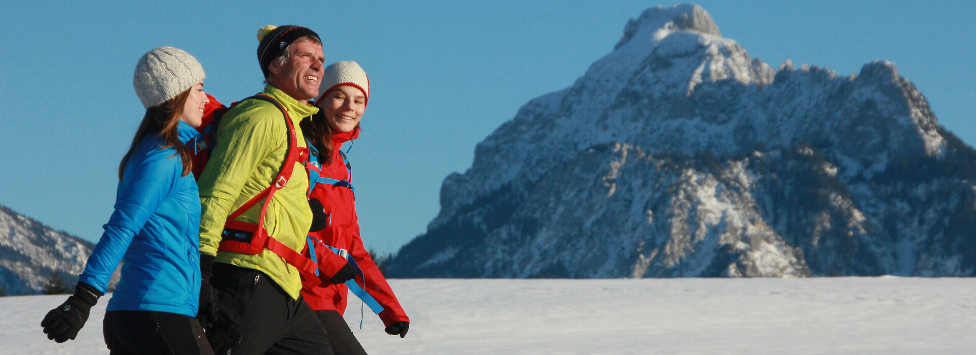 Das Bild zeigt zwei Frauen und einen Mann. Sie tragen Sport-Winterkleidung und laufen nebeneinander auf einem verschneiten Weg. Alle lächeln. Im Hintergrund sieht man die schneebedeckte Umgebung und die verschneiten Berge. Der Himmel ist blau. Die Sonne scheint.  