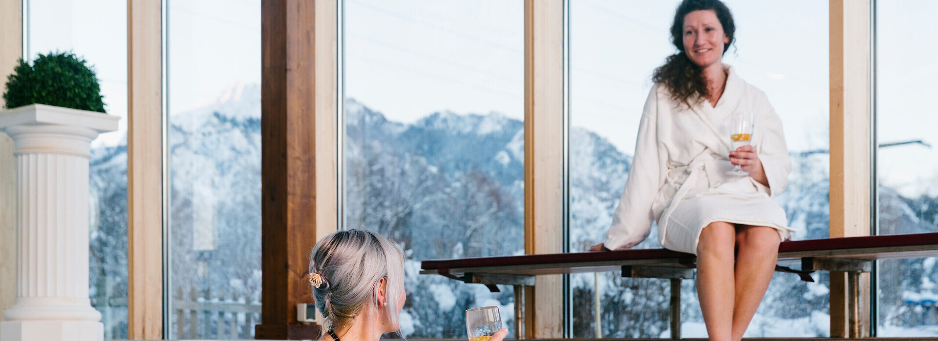 Das Bild zeigt einen Indoor-Swimming Pool. Eine Frau in Badekleidung ist im Pool und hält sich am Poolrand fest. Sie hält ein Wasserglas in der rechten Hand. Eine andere Frau sitzt auf einer Holzbank neben dem Pool. Diese Frau trägt einen weißen Bademantel. Auch sie hat eine gefülltes Wasserglas in der Hand. Die beiden Frauen lächeln sich zu. Hinter einer großen Fensterfront erheben sich die schneebedeckten Berge. Der Himmel ist grau.       