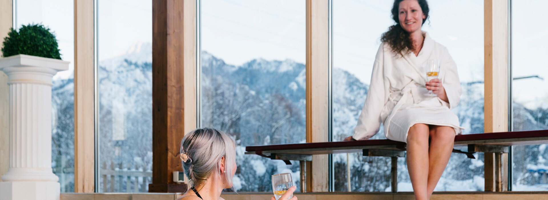 Das Bild zeigt einen Indoor-Swimming Pool. Eine Frau in Badekleidung ist im Pool und hält sich am Poolrand fest. Sie hält ein Wasserglas in der rechten Hand. Eine andere Frau sitzt auf einer Holzbank neben dem Pool. Diese Frau trägt einen weißen Bademantel. Auch sie hat eine gefülltes Wasserglas in der Hand. Die beiden Frauen lächeln sich zu. Hinter einer großen Fensterfront erheben sich die schneebedeckten Berge. Der Himmel ist grau.       