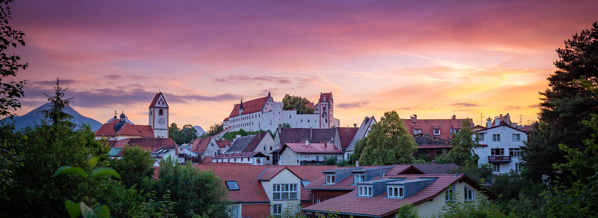  Das Bild fängt eine friedliche Sonnenuntergangsszene in Füssen ein. Der Himmel, der in Farbtönen von Lila und Pink gemalt ist, dient als schöne Kulisse für die Altstadt. Das Bild selbst ist ein charmanter Mix aus weißen und roten Gebäuden, deren Dächer eine Farbtupfer in die Szene bringen. Die allgemeine Atmosphäre des Bildes ist friedvoll und idyllisch und fängt das Wesen eines ruhigen kleinen Ortes in der Abenddämmerung ein.